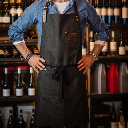 A person wearing a black denim apron with a leather strap and brass fittings. The apron has pockets and copper rivets, displayed in front of a backdrop of wine bottles.